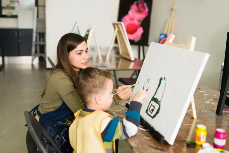 hispanic-female-teacher-is-instructing-caucasian-little-boy-use-brush-painting-blank-canvas-art-class